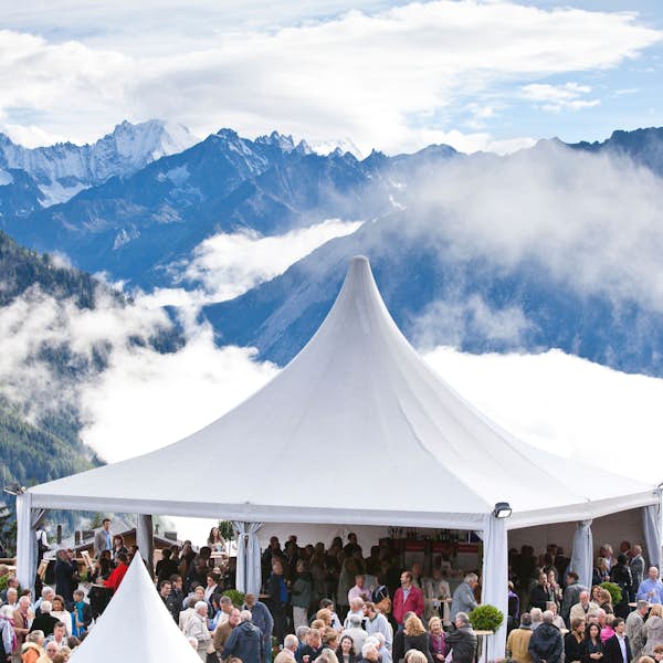 A crowd gathers under a large white tent set amidst a mountainous landscape with clouds and peaks in the background. A crowd gathers under a large white tent set amidst a mountainous landscape with clouds and peaks in the background.