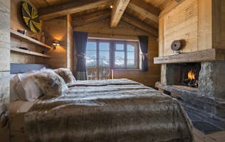 accommodation - Chalet Nyumba - Step into Chalet Nyumba, a cozy mountain retreat featuring a wooden cabin bedroom. Enjoy the large fur-covered bed, stone fireplace, and breathtaking window view of snowy mountains. Dark curtains frame the scene, enhancing the room's rustic and warm atmosphere.